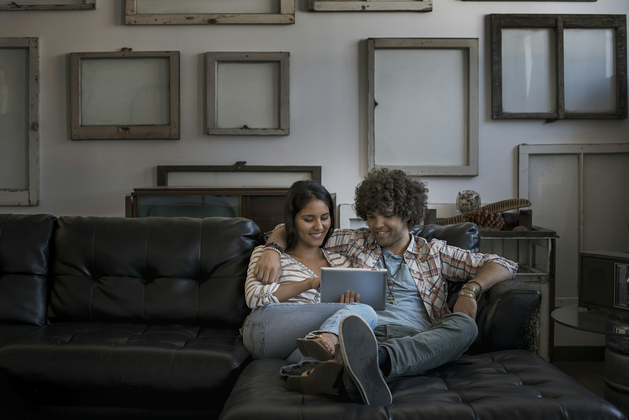 A couple on a sofa, looking at a digital tablet in front of a wall of empty picture frames.